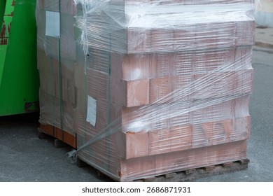 Stack of red building bricks tightly wrapped in transparent plastic stretch film standing on a wooden pallet at a construction site. Concept of renovation, industrial materials, and urban development.