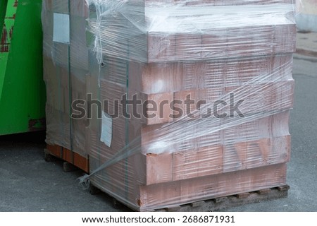 Stack of red building bricks tightly wrapped in transparent plastic stretch film standing on a wooden pallet at a construction site. Concept of renovation, industrial materials, and urban development.