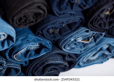 A stack of rolled jeans in assorted blue shades and black, arranged neatly on a white background, showcasing casual fashion and textile variety in an organized display
