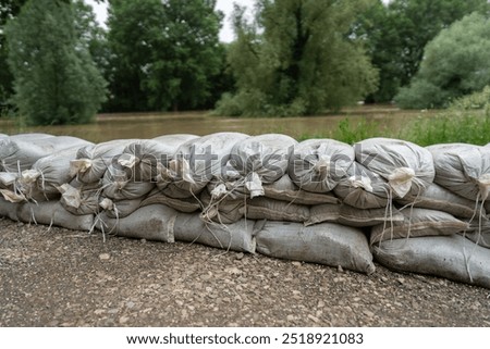 stack of sandbags forms a temporary flood barrier along a riverbank, protecting against rising floodwaters. The sandbags are neatly arranged to safeguard the area from flooding
