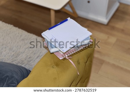 Stack of school notebooks with word HOMEWORK on top page and blue pen on mustard yellow upholstered furniture