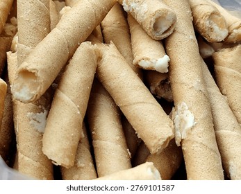 A stack of semprong, or egg rolls, fills the entire frame. The cakes are thin, light brown tubes with a finely porous surface and the characteristic spiral lines of the rolls. Many of the ends are broken, revealing a soft white cream filling inside. A few crumbs of cream also cling to the outside. The texture is crumbly, light, and crisp. The natural lighting highlights the details of the pores in the cake crust and the contrast between the dry crust and the creamy cream.
