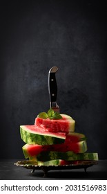 a stack of slices of ripe red watermelon in a metal plate, stuck in a large knife. Black background