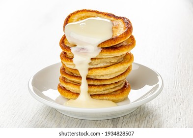 Stack Of Small Pancakes Poured Condensed Milk In White Saucer On Wooden Table