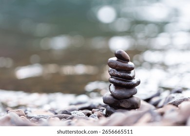 Stack of smooth black pebbles balanced on a rocky riverbank with blurred water background. Represents balance, meditation, harmony, nature, and mindfulness in a peaceful outdoor setting.