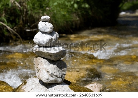 A stack of smooth, carefully balanced stones stands on the bank of a clear stream. Sunlight highlights the water flowing gently past in a tranquil outdoor setting.