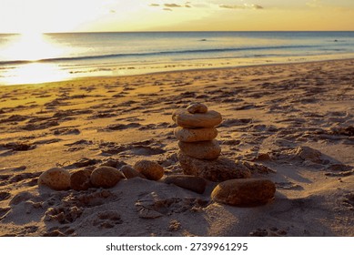 Stack of smooth stones balanced on sandy beach at sunset with warm golden light reflecting over calm ocean waves and peaceful horizon.