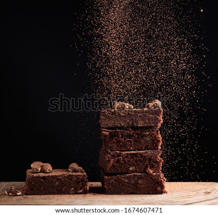 stack of square baked brownie pieces sprinkled with cocoa powder, particles froze in the air against a dark background, delicious chocolate dessert