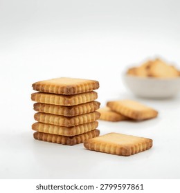A stack of square shortbread cookies on a white surface with a few cookies scattered around and a bowl of cookies in the background.