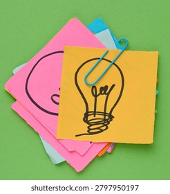 Stack of sticky notes with a light bulb drawn on them is held together with a blue paper clip against a green background. 
