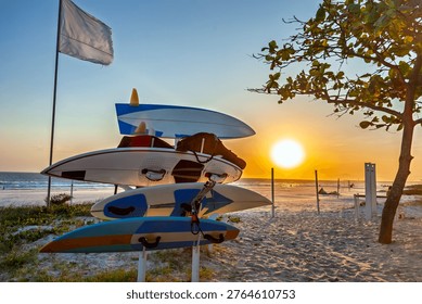 A stack of surfboards on a rack at Praia do Pepê, Rio de Janeiro, during a golden sunset. The beach scene features white sand, a flag, and a tree, capturing the vibrant kitesurfing culture.
