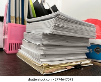 A stack of thick, slightly worn documents sits on a wooden desk, with edges uneven from frequent use. The papers are piled high, suggesting heavy workload or extensive records. A binder clip holds some pages together at the bottom, while a pink file holder and several books stand in the background. The scene conveys a busy office or study environment, with paperwork as the main focus.