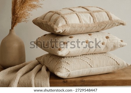 A stack of three neutral-colored throw pillows with different textured designs. The top pillow features a grid pattern, the middle pillow has small dot embellishments, and the bottom pillow showcases a braided design. The pillows are placed on a wooden surface with a beige blanket and a vase with dried grass in the background.