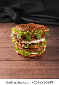 Stack of three simit sandwiches top of each other on wooden table and black  background. Round bagel sandwiches with different ingredients. Mint leaves, white cheese, tomatoes, ham, lettuce.