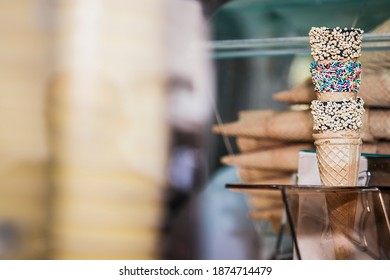 Stack of three waffle ice-cream cones decorated with nuts, sprinkles and chocolate glaze, stacked in cone holder on counter of ice-cream parlour