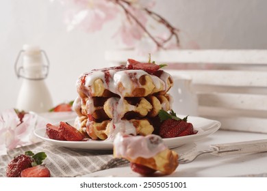 stack of three waffles on a white plate, with a generous drizzle of white icing and strawberry sauce on top with fresh strawberries are scattered around the plate, and a sprig of cherry blossoms . - Powered by Shutterstock