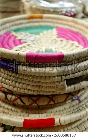 Stack of traditional handwoven baskets shows natural straw fibers with colorful thread accents in pink, red, and purple. The circular trays are piled neatly, highlighting craftsmanship and texture 
