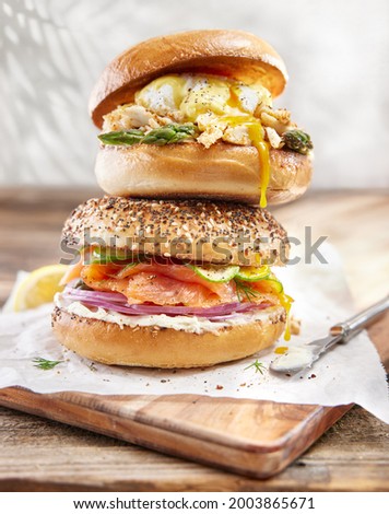 A stack of two bagels. Bottom is an everything bagel with lox, cream cheese, red onion and sliced cucumbers.  Top is a poached egg with hollandaise sauce and asparagus.  Egg yolk is dripping down. 