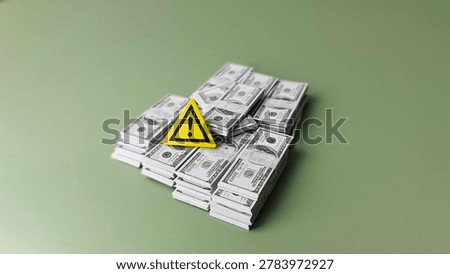 Stack of US dollar banknotes with yellow warning triangle icon on green background symbolizing financial risk and loss. 