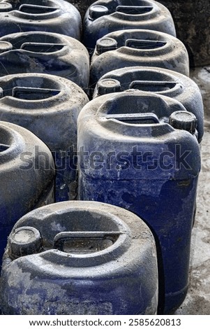 Stack of used blue plastic jerrycans arranged on a concrete floor, showing signs of dirt and wear. Commonly used for storing and transporting liquids such as chemicals, fuel, and industrial fluids.

