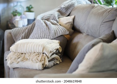 Stack of a variety of soft knit throw blankets stacked on a grey couch in a farmhouse style living room. Selective focus on covers with blurred foreground and background.