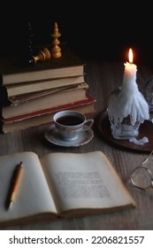 Stack of vintage books, cup of tea or coffee, lit candles, reading glasses and chess pieces on wooden table. Dark academia concept. Selective focus.