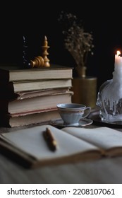 Stack of vintage books, cup of tea or coffee, lit candles, reading glasses and chess pieces on wooden table. Dark academia concept. Selective focus.