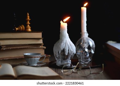 Stack of vintage books, cup of tea or coffee, lit candles, reading glasses and chess pieces on wooden table. Dark academia concept. Selective focus.