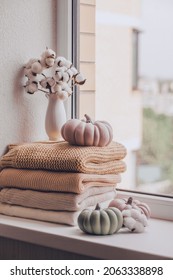 A Stack Of Warm Knitted Sweaters On The Windowsill With Pumpkins And A Vase Of Cotton Plant On A Rainy Day, In Pastel Shades.