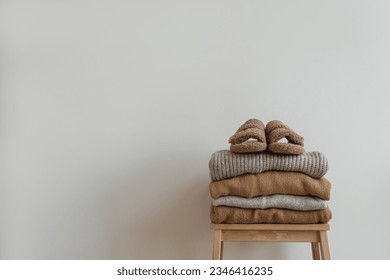 Stack of warm neutral beige clothes on wooden stool over white wall. Fluffy slippers, wool pullovers and sweaters. Minimalist autumn, fall fashion background