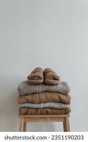 Stack of warm neutral beige clothes on wooden stool over white wall. Fluffy slippers, woolen pullovers and sweaters