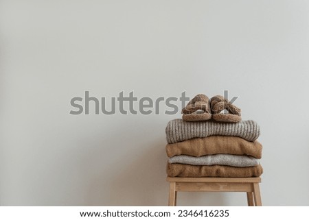 Stack of warm neutral beige clothes on wooden stool over white wall. Fluffy slippers, wool pullovers and sweaters. Minimalist autumn, fall fashion background