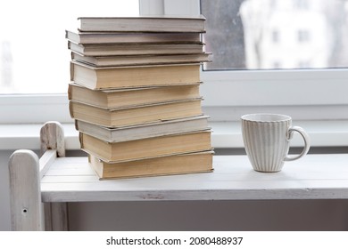 A stack of white-bound books is on the table against the background of the window. The cup of tea is next to books. School education concept.