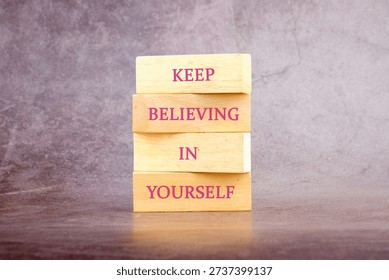 Stack of wooden blocks displaying Keep Believing In Yourself on a dark surface with a grey wall background.