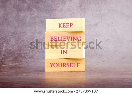Stack of wooden blocks displaying Keep Believing In Yourself on a dark surface with a grey wall background.