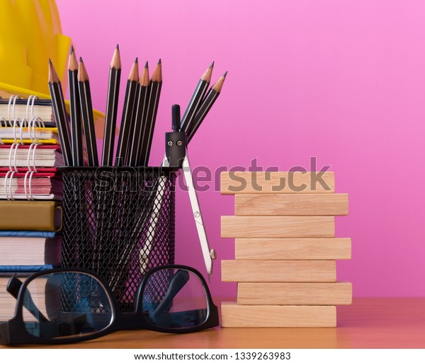 Stack wooden blocks with\
pencils, dividers, glasses, construction helmet and stack of book\
on wooden table isolated pick background, Blank wooden blocks for\
your text.