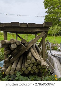 A Stack Of Wooden Posts Sat Under A Wooden Table Outdoors In A Garden Setting With Cloudy Sky Grass And Trees With An Upside Down Wheelbarrow Off To The Side