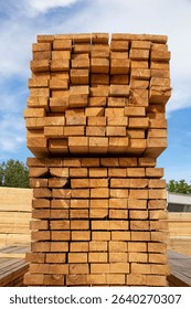Stack of Wooden Timber Beams at Lumber Yard
					A large stack of square-cut wooden beams arranged neatly at an outdoor lumber yard. The timber is stored on a gravel surface and appears seaso