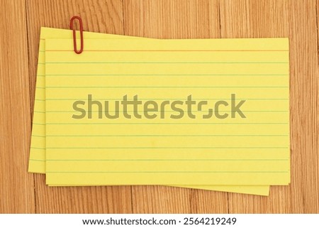 A stack of yellow lined index cards paper held together with a red paperclip on a wooden desk, perfect for notes, reminders, and organization.