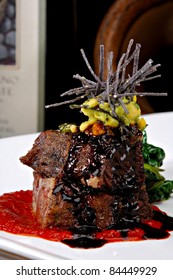 Stacked Braised Short Ribs On A Bright Red Tomato Paste Sauce.  Garnished With Avocado, Sliced Tortilla Chips And A Red Wine Reduction Sauce.  Colorful Sauteed Spinach Is Displayed Behind The Meat.