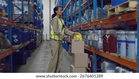 Stacking boxes worker wearing safety vest in storage aisle with hazard drums and yellow helmet. Industrial, logistics, safety, organization, storage, inventory, operational