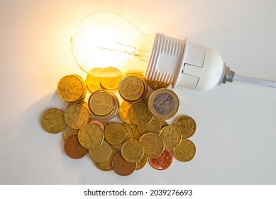 Stacks Of Euro Coins With Lit Light Bulb Beside, Placed On White Wooden Surface. Value Of Money And Energy Tariff Trends. 
