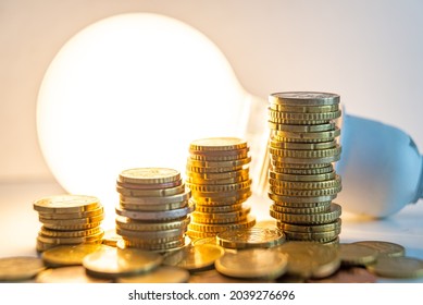 Stacks Of Euro Coins With Lit Light Bulb Beside, Placed On White Wooden Surface. Value Of Money And Energy Tariff Trends. 
