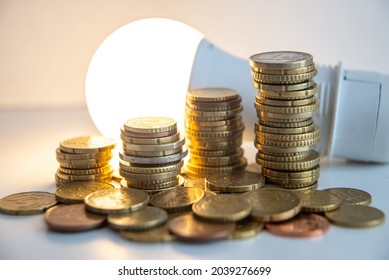 Stacks Of Euro Coins With Lit Light Bulb Beside, Placed On White Wooden Surface. Value Of Money And Energy Tariff Trends. 
