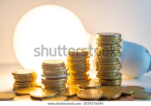 Stacks of euro coins with lit light bulb beside,\
placed on white wooden surface. Value of money and energy tariff\
trends. \
