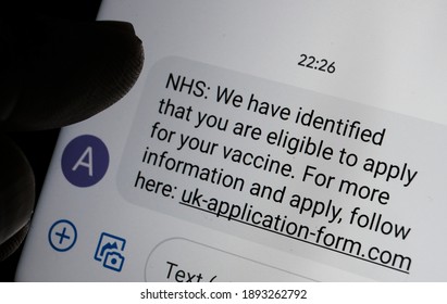 Stafford, United Kingdom - January 13 2021: Scam coronavirus vaccine text message seen on the smartphone screen and blurred silhouette of finger pointing at it. 