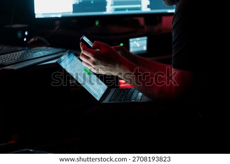 Stage engineer managing digital tools on laptop and smartphone under low light conditions in backstage control environment during technical workflow