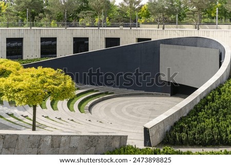 Stage with large screen is located in front of wide semicircular benches for spectators. Mature oak trees grow on terraces of amphitheater. City park "Krasnodar" or "Galitsky Park". Sunny spring 2024