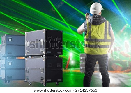 Stage logistics supervisor coordinates equipment movement via radio, managing technical gear and ensuring smooth setup during an active laser-lit event preparation.