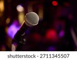 Stage microphone close-up ready for performance with shallow bokeh background, textured grille catching warm stage LEDs and blurred band equipment suggesting